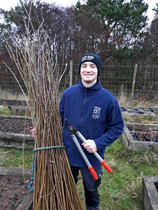 A young man brandishing a pair of loppers and cradling a bundle of reeds.