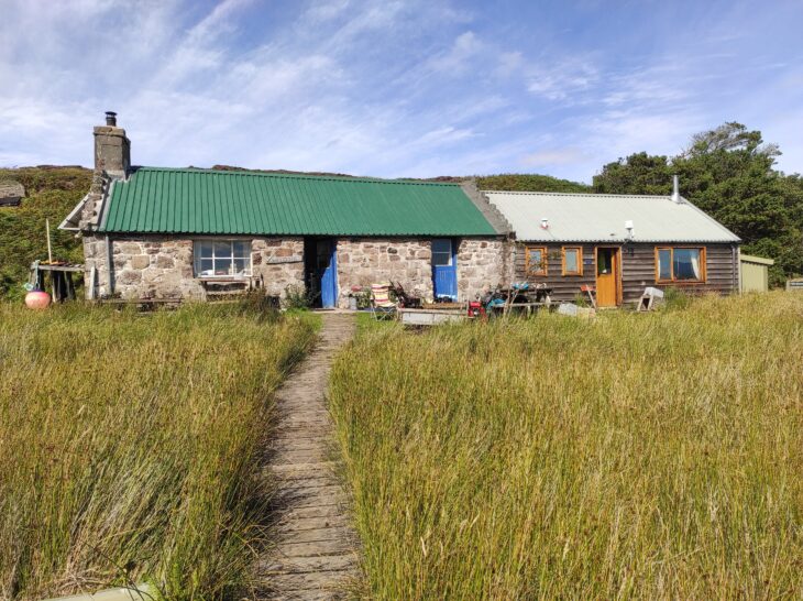 A modest stone building with a green roof and timber extension sits in a field of long grass with a rough path leading to its front door.