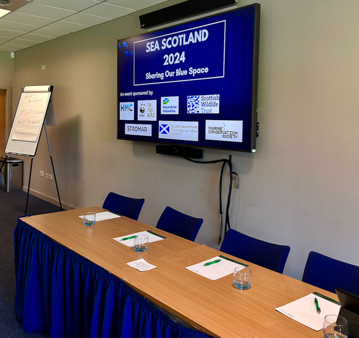 A table with notepads and pens with a screen behind displaying the Sea Scotland 2024 logo.