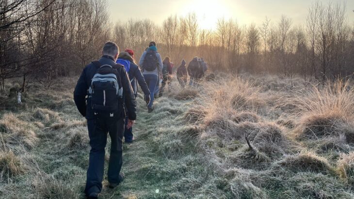 A group of people walk through long grass on a frosty morning. In the background the sun is shining through trees that have lost their leaves in winter.