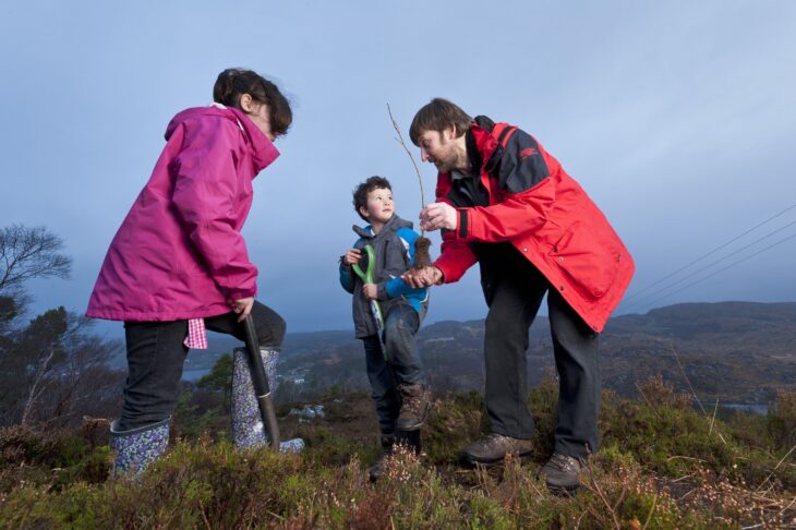 Three people are planting a tree. A woman is digging a hole while a man holds a tree sapling out to a child.