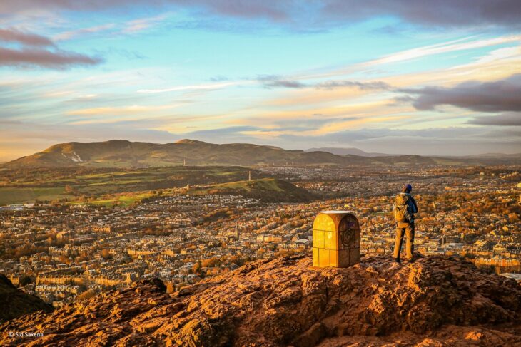 A person standing on top of a hill, next to a trig point, looking over a city. 