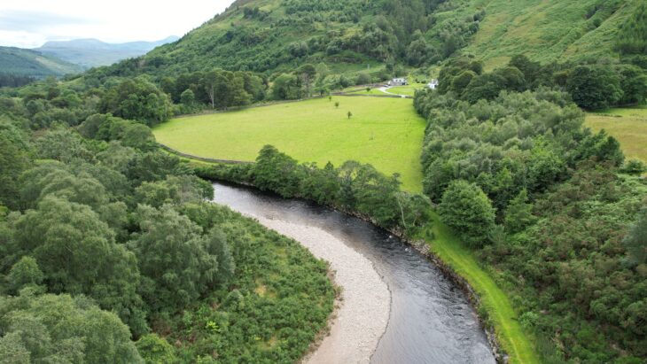 A bending river surrounded by woodlands and a grassy field.