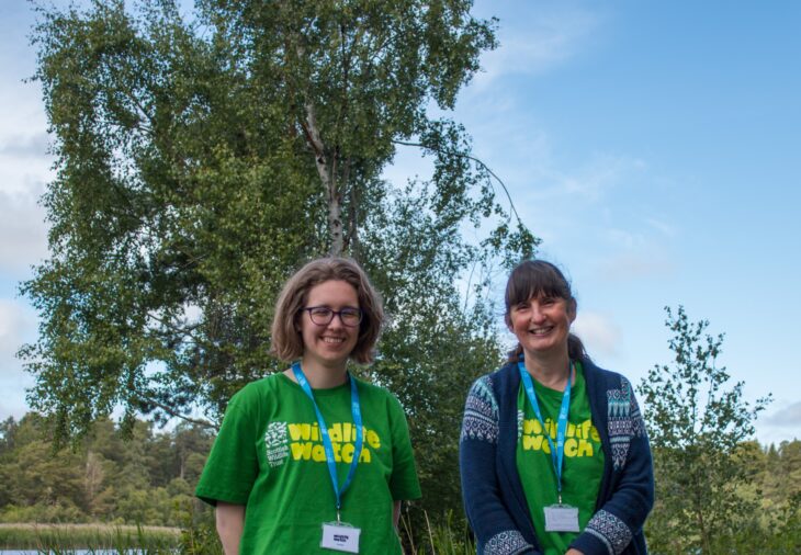 Two people wearing green Wildlife Watch t-shirts are standing in front of a beech tree.