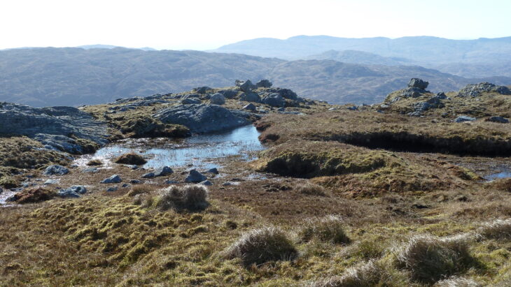 A small lochan amongst tussocks of long grass and rocky outcrops. In the background are the hills of the Scottish Wildlife Trust's Rahoy Hills reserve.