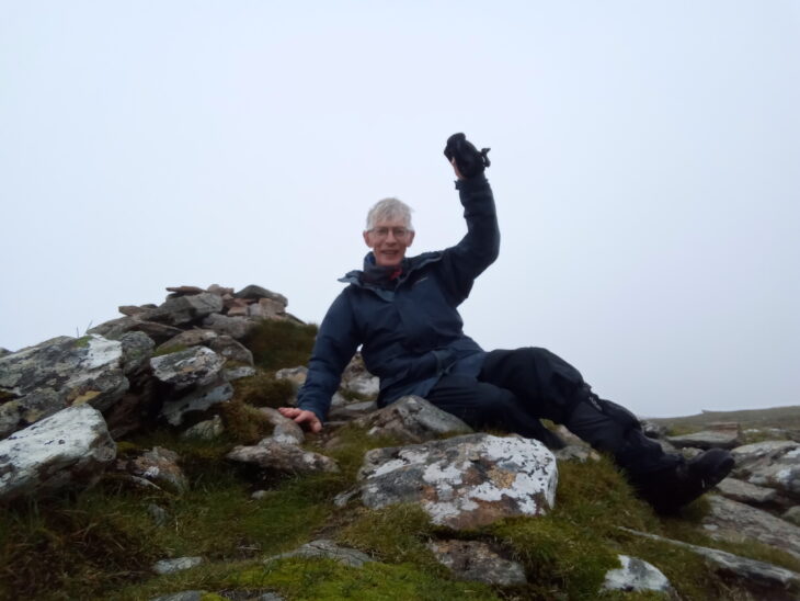 A photo of a man laying on a small pile of rocks. The man is wearing a waterproof coat and walking clothes and is smiling and waving towards the camera. Behind him is a thick bank of fog obscuring the mountain view.Photo taken by John Knox.