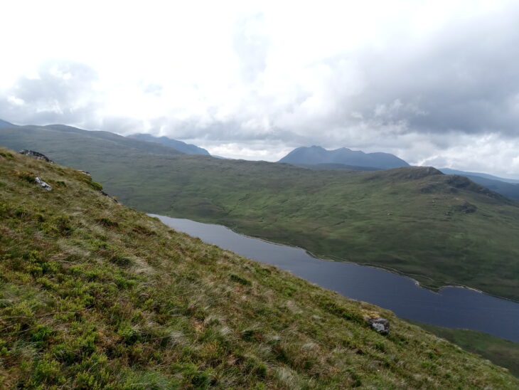 A photo showing mountain views. In the foreground there is a grassy slope where the photo is taken from. Beyond is a long loch that is dark blue. In the distance are more hills and mountains shrouded in clouds.Photo taken by John Knox.