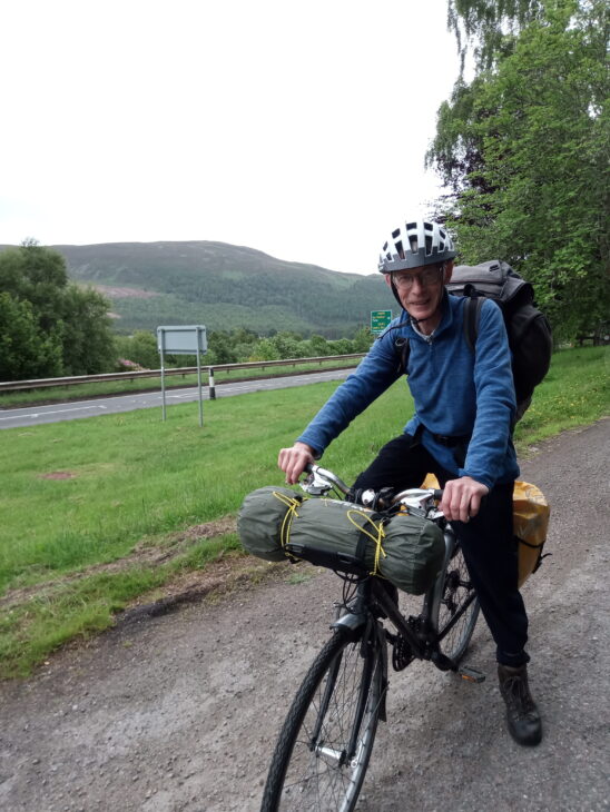 A photo of a man on a bicycle. The man has a cycling helmet and a rucksack on and there is a tent strapped to the front of the bike.Photo taken by John Knox.