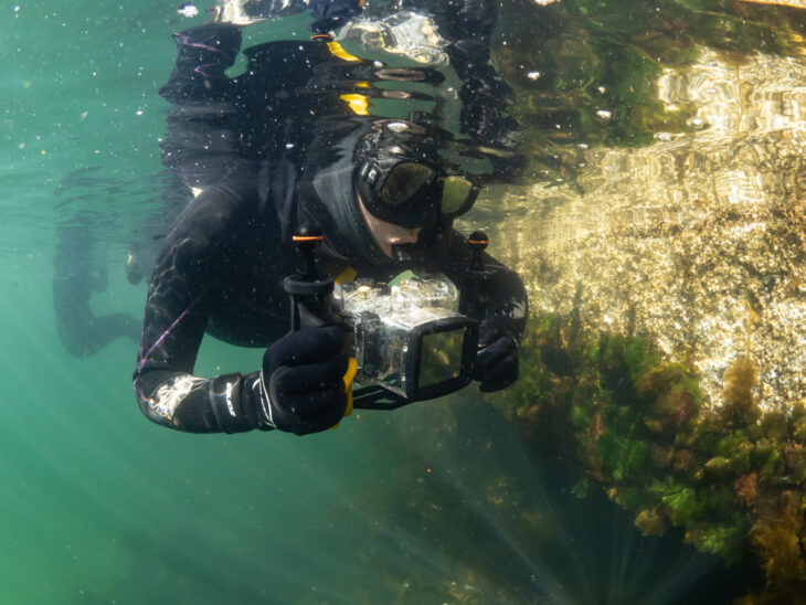 A snorkeller swimming by underwater rocks, holding a camera.