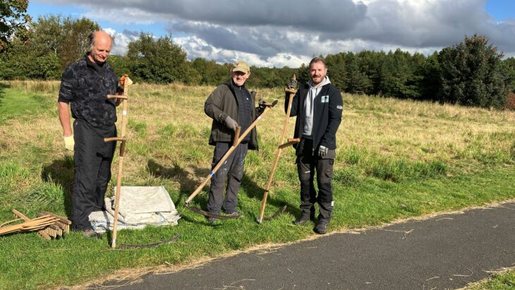 Three volunteers holding scythes on a patch of grass beside a footpath.