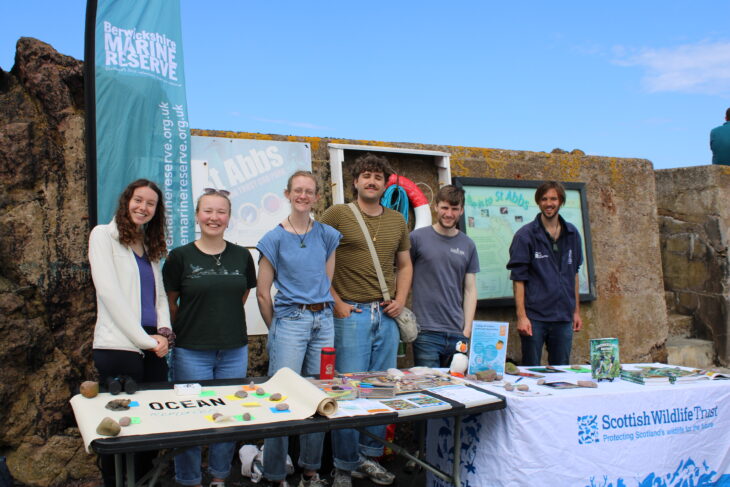 Six Young Leaders standing side by side at a table with various leaflets, posters and books about snorkelling and the Scottish Wildlife Trust.