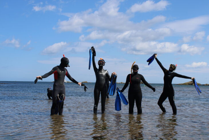 Four people wearing snorkels and wetsuits are standing in shallow water, holding pairs of flippers.